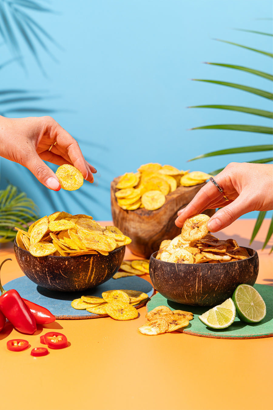 Two hands reaching into bowls of plantain chips with fresh chillies and lime